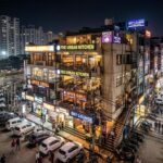 4K photograph of a crowded, narrow street in Hajipur, Sector 104, Noida, lined with multi-story cafés and restaurants operating in abadi lands without fire safety compliance.